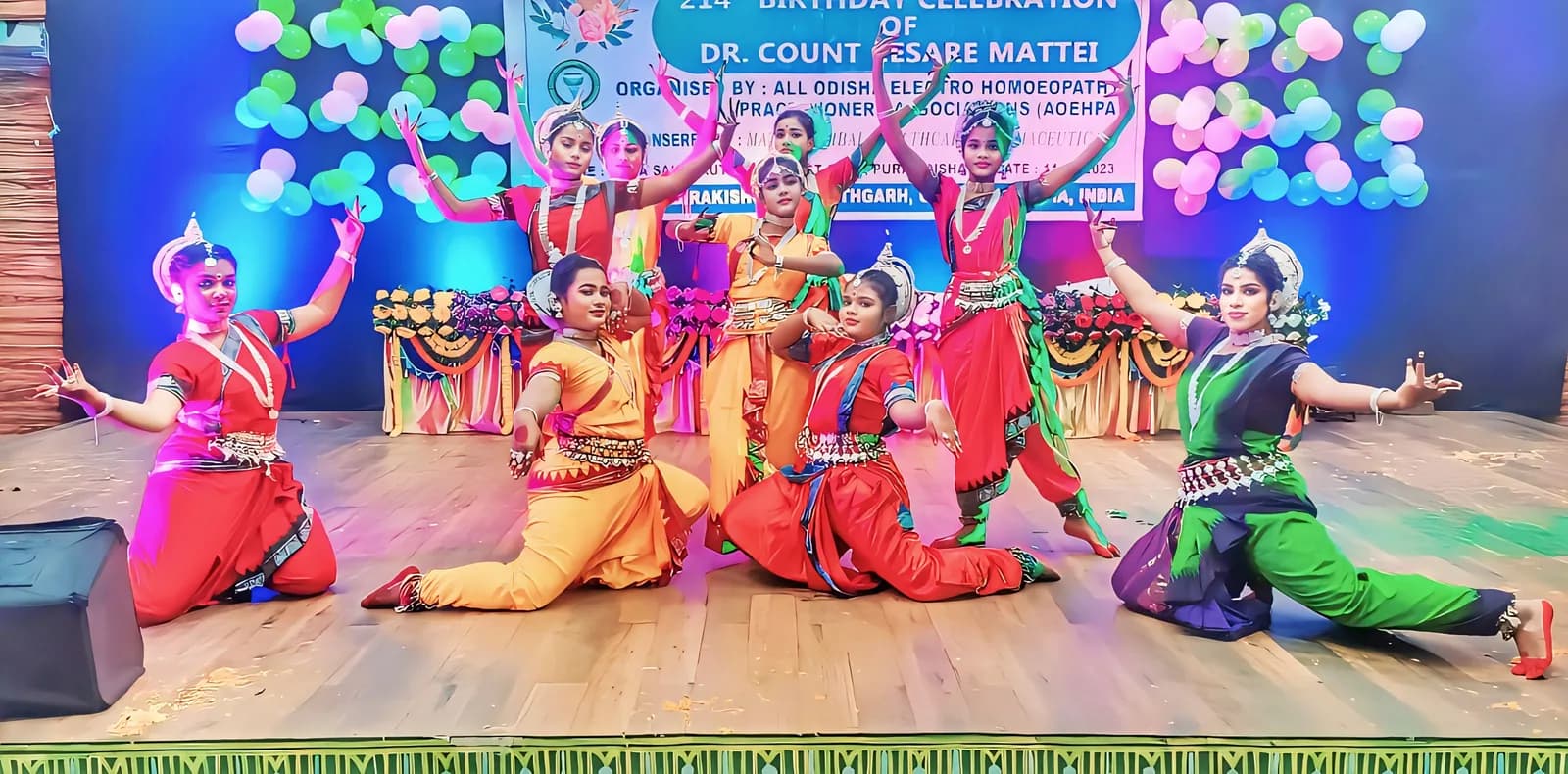 Nine dancers in kneeling and standing Odissi poses across a brightly lit stage